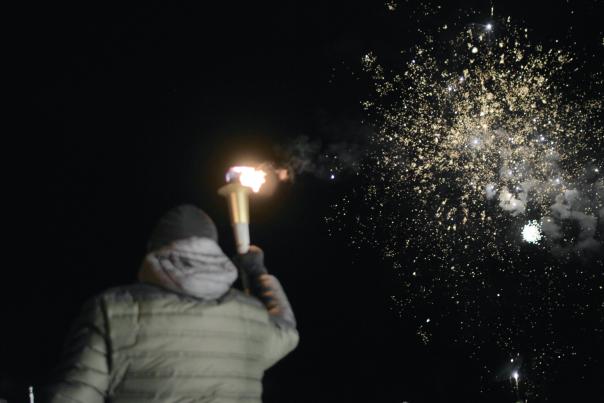 A man holding a lit torch to the dark sky with golden fireworks overhead at the Opening Ceremony of hg6668皇冠登录's Meijer Winter State Games.