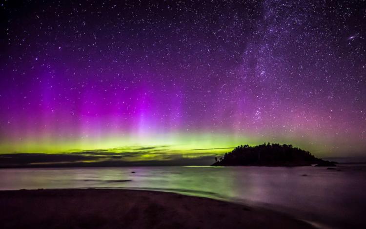 A purple and green Aurora Borealis reflecting off of the waters with stars in the sky
