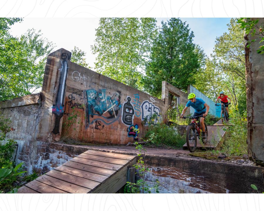 Two bikers riding down wooden bridges between concrete walls tagged with graffiti