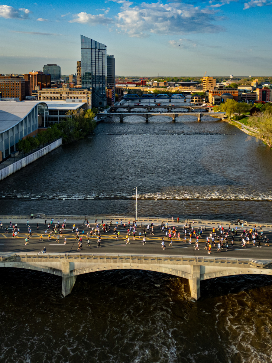 A wide river flows through a city with a marathon in progress on a bridge in the foreground and a skyline of buildings in the background.