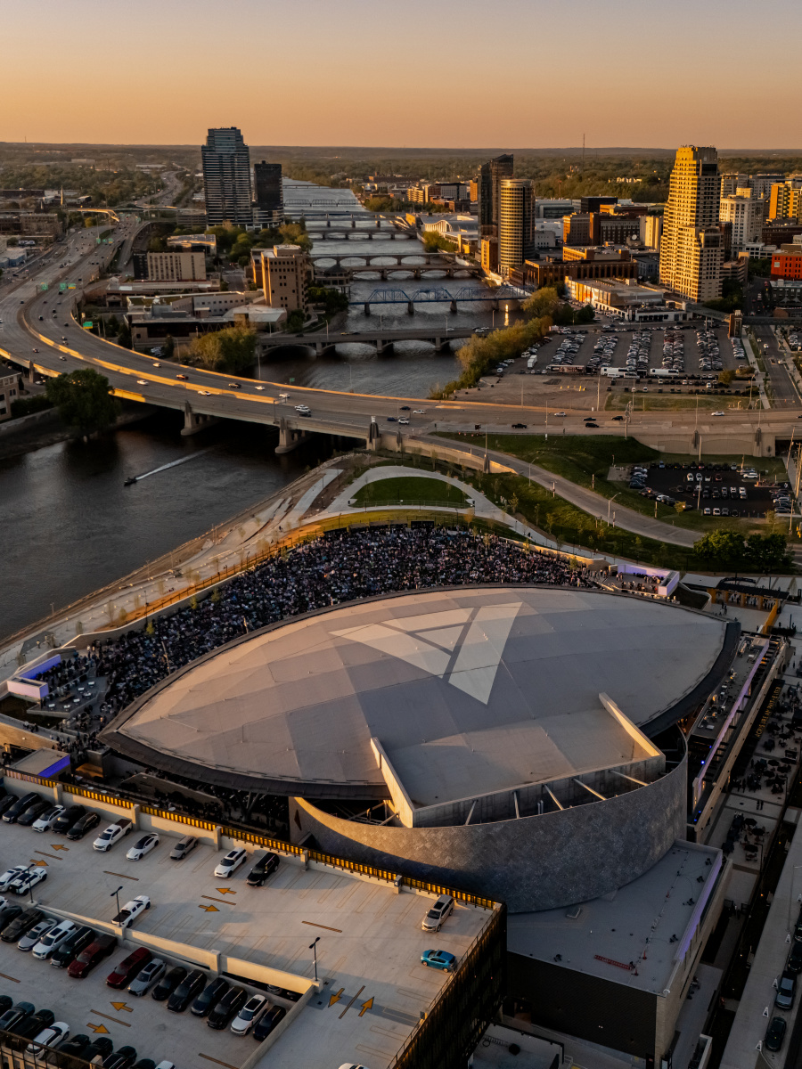 A large, modern amphitheater filled with people sits beside the Grand River in the foreground, while the Grand Rapids city skyline with bridges and highways stretches into the distance under a warm sunset.