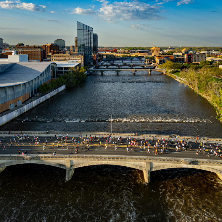 A wide river flows through a city with a marathon in progress on a bridge in the foreground and a skyline of buildings in the background.