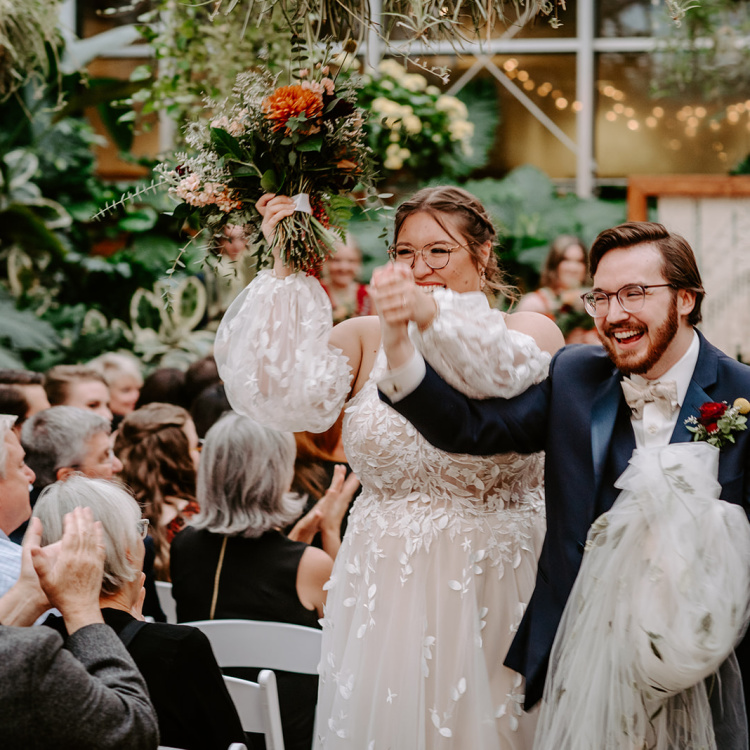 A joyful bride and groom celebrate their marriage in the foreground, surrounded by seated guests in a lush, greenhouse-like setting filled with greenery.