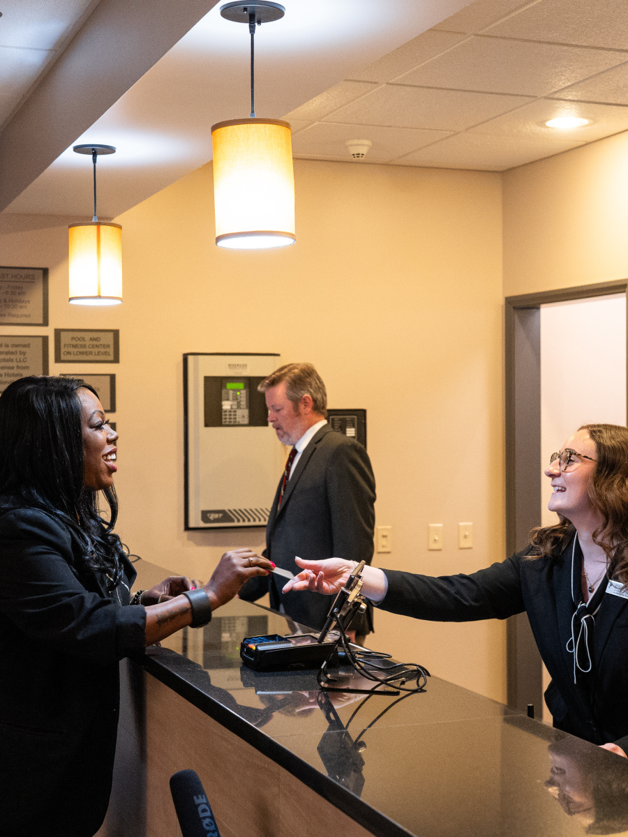 The image shows three people in a professional setting, with two women shaking hands and a man standing nearby.