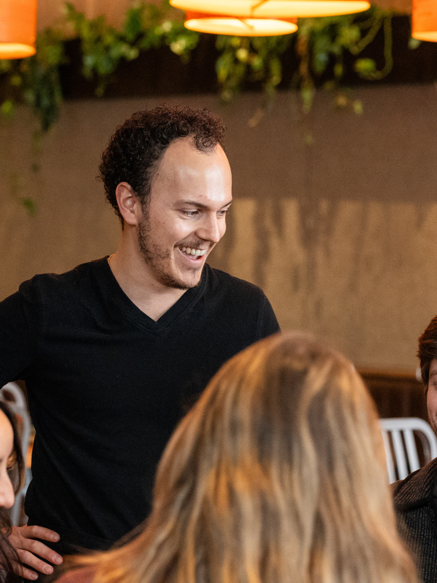 A group of people, including two men and two women, are engaged in a lively conversation in a cozy, dimly lit restaurant setting with warm lighting and greenery in the background.