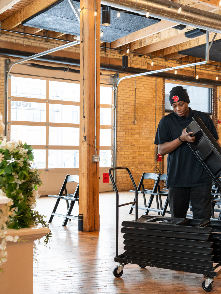 A person in black clothing is standing in a spacious room with wooden beams and large windows, surrounded by chairs and a floral arrangement.