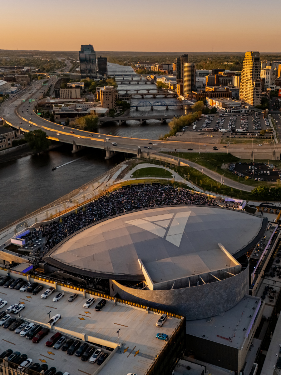 A large, modern amphitheater filled with people sits beside the Grand River in the foreground, while the Grand Rapids city skyline with bridges and highways stretches into the distance under a warm sunset.