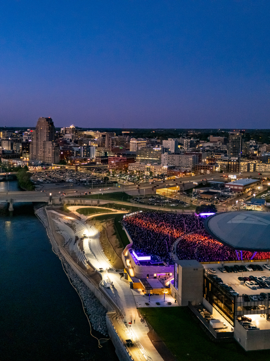 A river flows past the Acrisure Amphitheater filled with spectators in the foreground, with the Grand Rapids city skyline illuminated by lights in the background under a twilight sky.