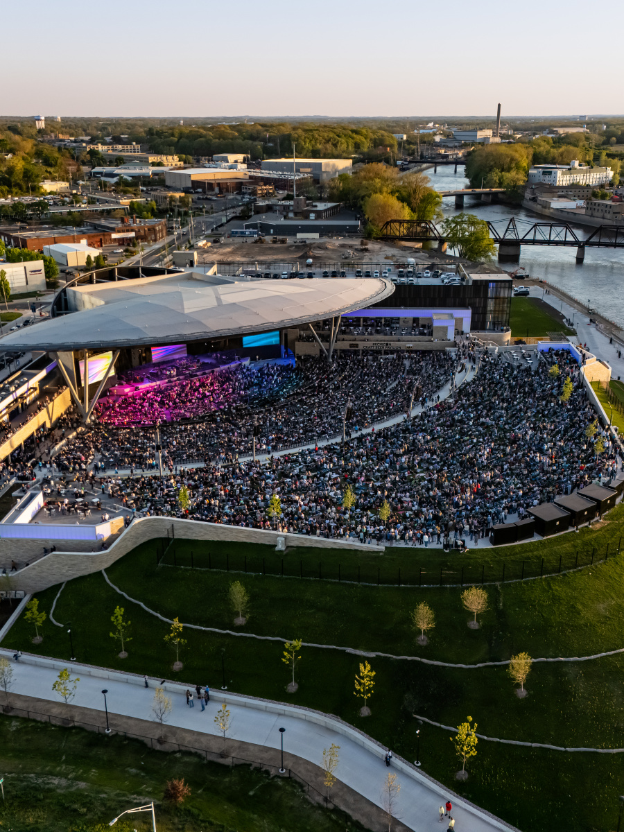 The Acrisure Amphitheater outdoor concert venue filled with a crowd is situated in the foreground, with a river and cityscape visible in the background.