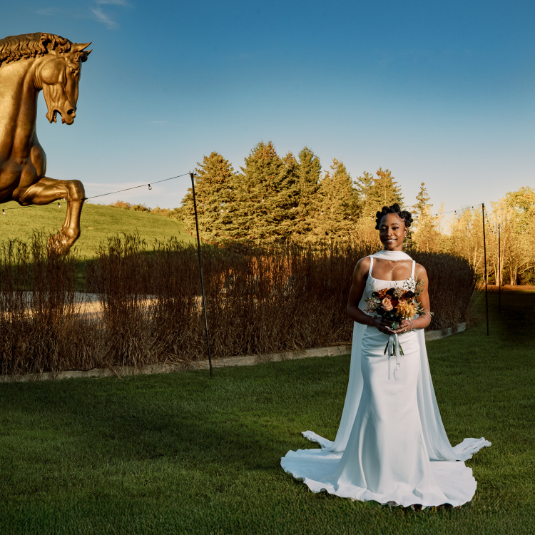 A bride in a white gown holds a bouquet in the foreground, with a large golden horse statue and a line of autumn trees in the background.