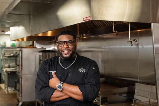 A smiling man wearing a black chef's uniform stands in a commercial kitchen, with stainless steel appliances and equipment visible in the background.