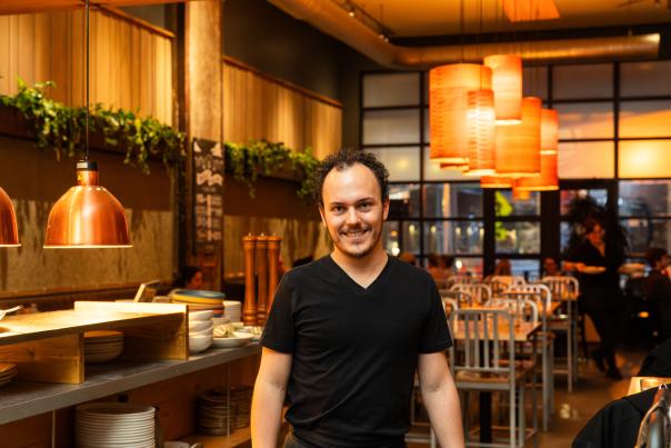 A man in a black shirt stands in a cozy, dimly lit restaurant setting with wooden furniture, hanging plants, and warm lighting.