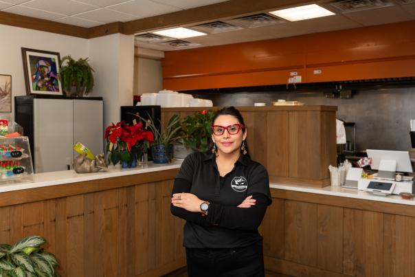 The image shows a smiling woman wearing a black sweater and glasses, standing in what appears to be a cafe or restaurant setting with various decor and furnishings visible in the background.