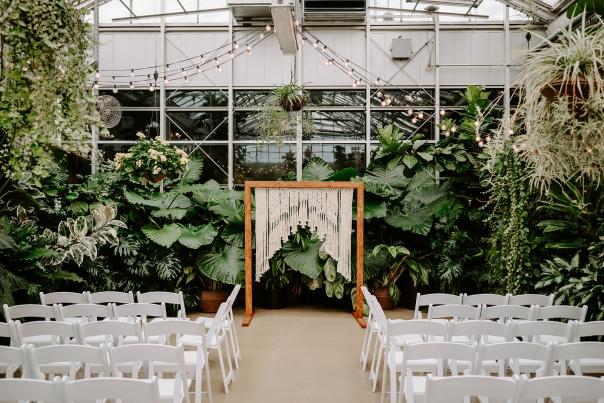 Rows of white folding chairs face a macramé wedding arch set against a lush backdrop of tropical plants inside a glass greenhouse.