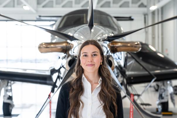 A woman with long brown hair stands in front of a large propeller-driven aircraft in a hangar.