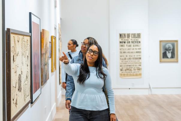 The image shows a group of people examining artwork displayed on the walls of the Grand Rapids African American Museum & Archives. The foreground features a woman in a light-colored sweater standing in front of the artwork, while the background includes additional framed pieces and what looks like a text-based display.