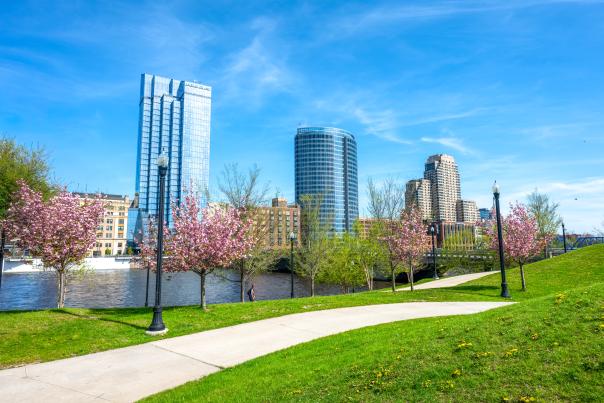 A winding path through a grassy park with blooming cherry trees leads to a city skyline of modern skyscrapers under a bright blue sky.