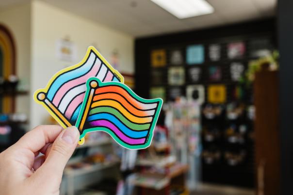 A hand holds two pride flags in the foreground, with a blurred background of shelves filled with items.