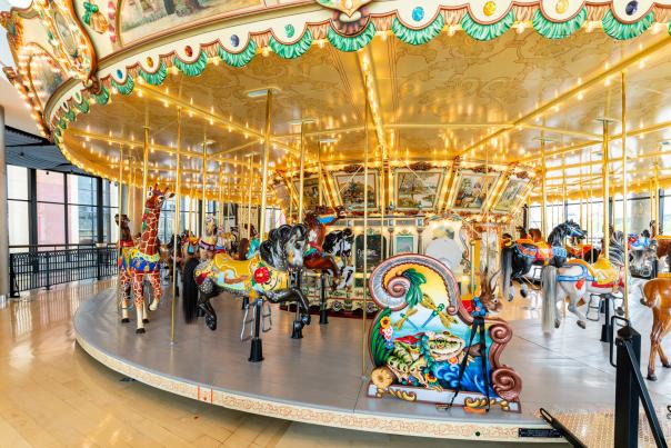 A brightly lit carousel with various animal figures in the foreground is set against a backdrop of large windows and a ceiling with decorative trim.