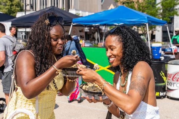 Two women in the foreground enjoy food from black bowls, while in the background, tents and a Jamaican flag suggest an outdoor festival.