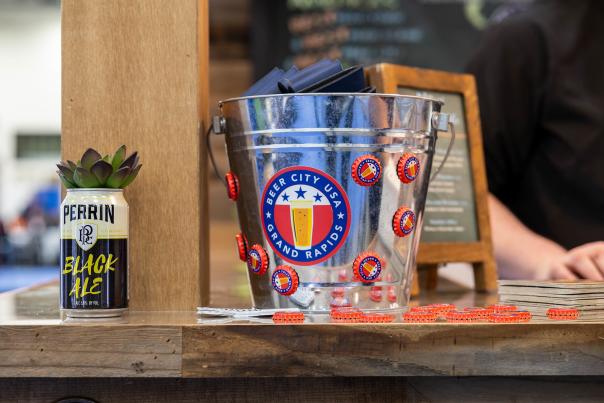 A metal bucket adorned with bottle caps and a "Beer City USA" sticker sits on a wooden counter next to a can of Perrin Black Ale with a succulent, while a person's hands are visible in the blurred background.