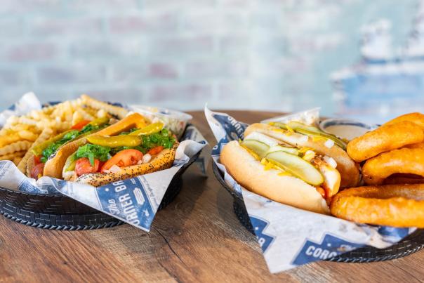 Two baskets of food, one with hot dogs and onion rings and the other with a hot dog and fries, sit on a wooden table in front of a brick wall.