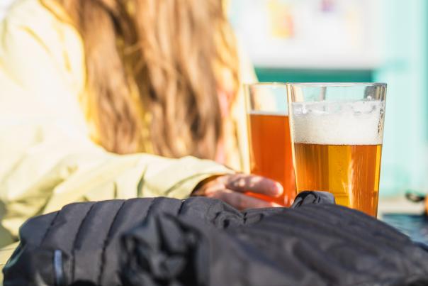 Two glasses of beer sit on a table in the foreground, with a woman in a yellow jacket and beanie blurred in the background in front of a teal building.
