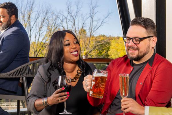 A group of people enjoying drinks and conversation on an outdoor patio, surrounded by a scenic natural setting with trees and a clear sky.