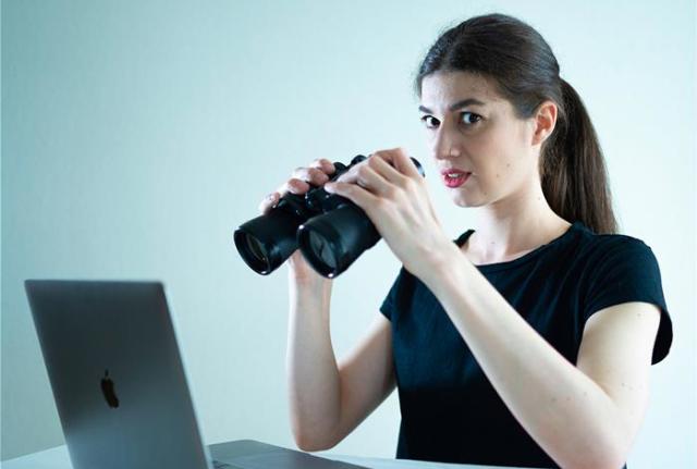Image of lady siting at a desk with a laptop and using binoculars