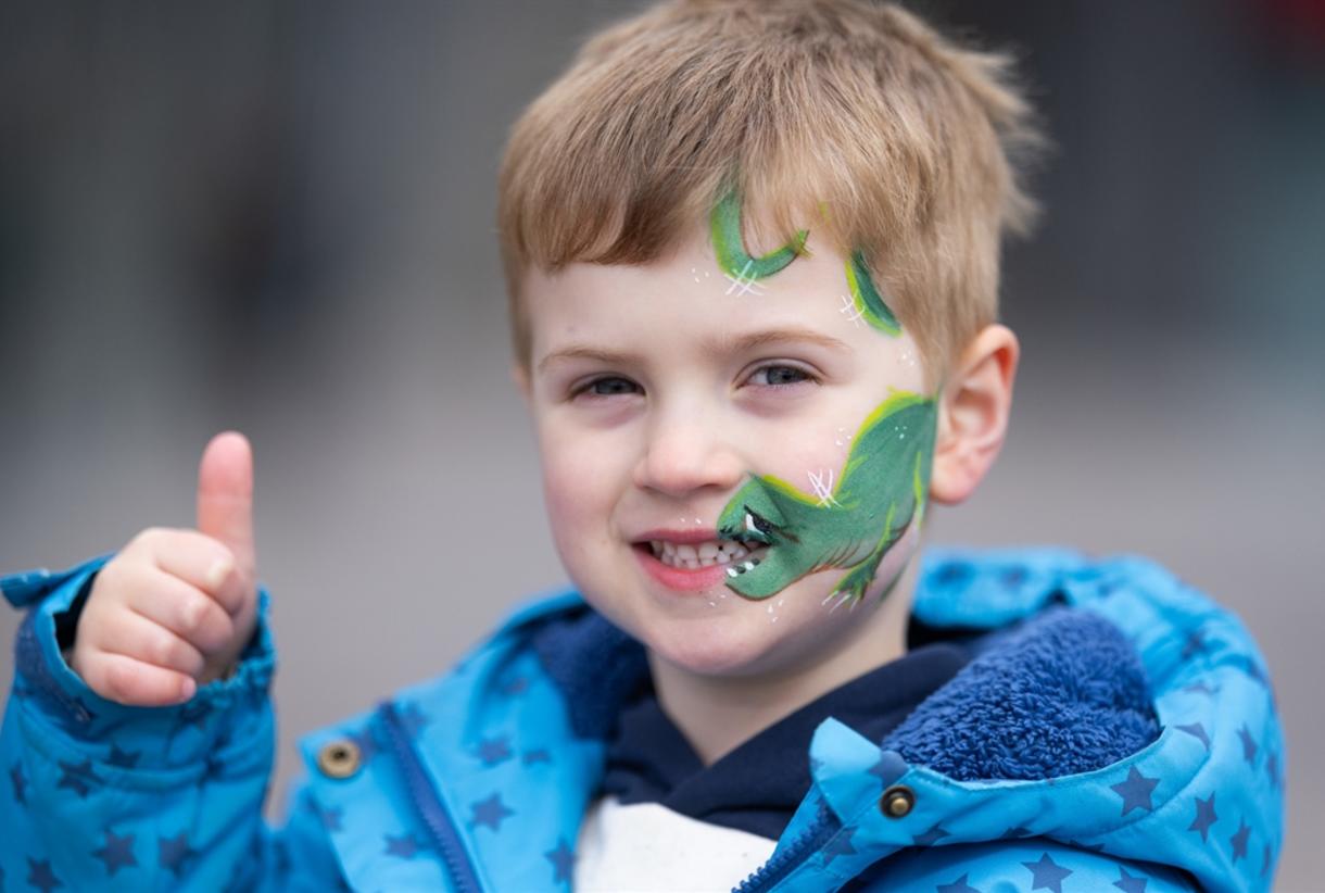 A young child wearing a blue jacket gives a thumbs up while smiling at the camera, with a green dino…