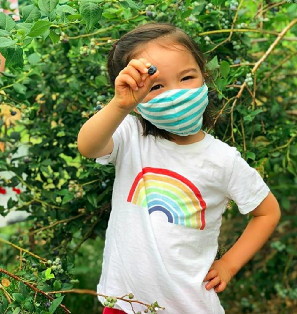 A masked child holding up a blueberry picked at Terhune Orchards near Princeton