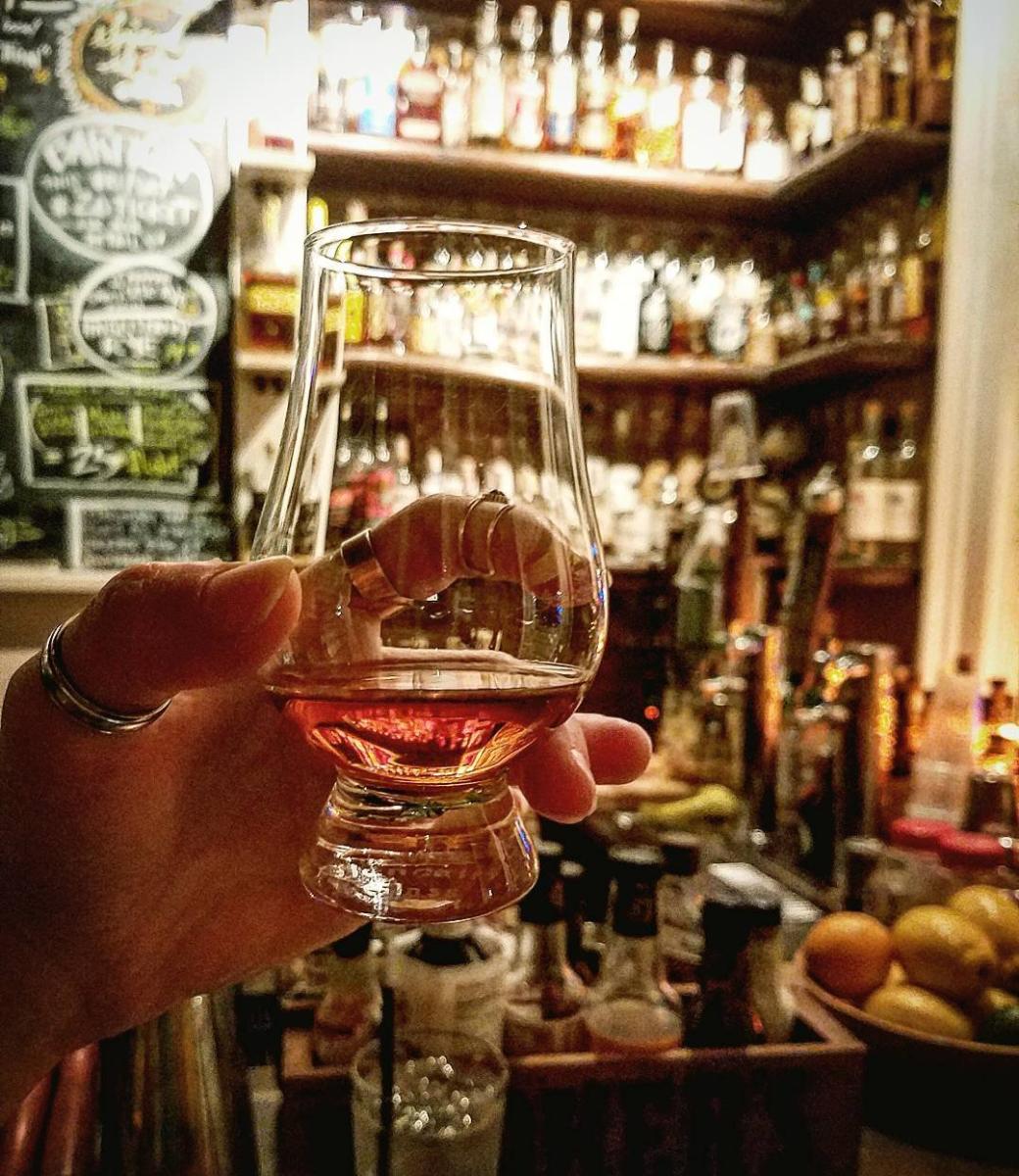 Glencairn glass of bourbon held up with Old Kentucky Bourbon Bar's huge bourbon collection behind it.