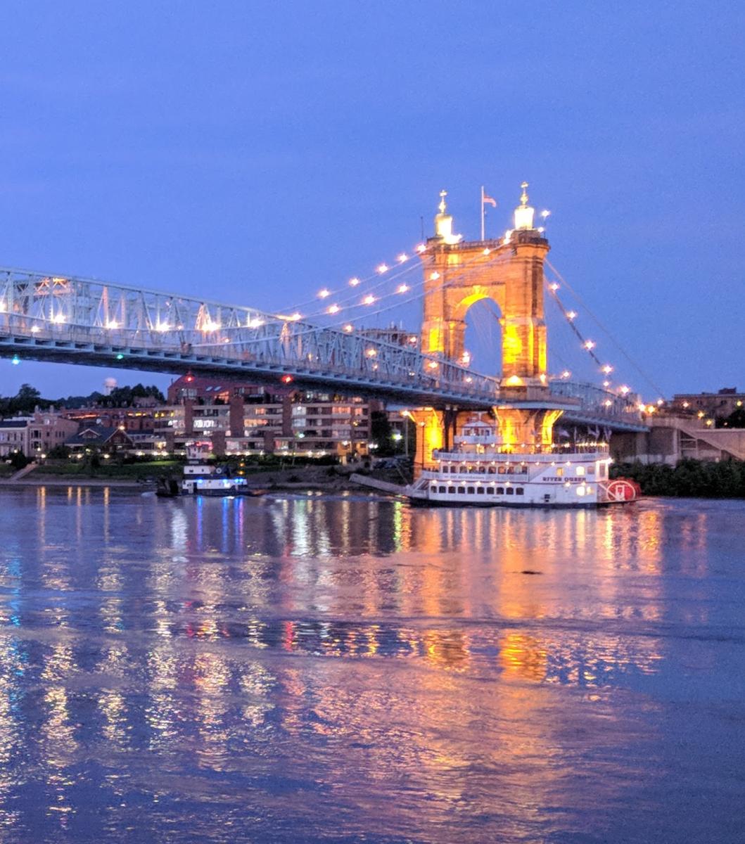 The Roebling Suspension bridge connecting Northern Kentucky to Cincinnati, with the Ohio river looking pink and blue in the sunset and a BB Riverboat sailing under the bridge