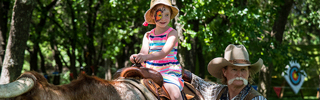 Child riding long horn cow with help from a cowboy