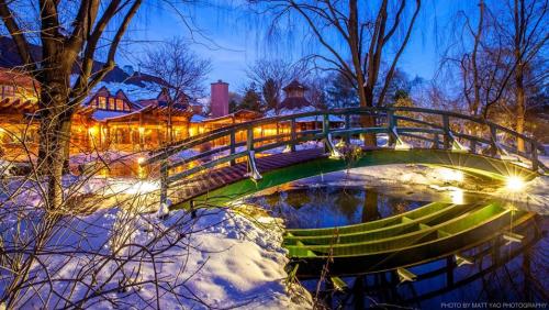 A view of Rat's restaurant from across a pond with snow and a walking bridge lit up at dusk