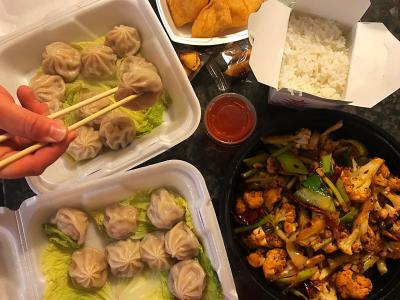 Bird's-eye view of Chinese dinner with dumplings, stir fry, and rice in take-out boxes. Hand holding chopsticks grabbing dumpling in corner.