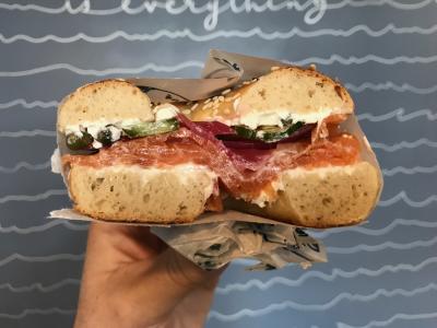 Hand holding half of bagel sandwich topped with meat, greens and spread from Lox Bagel Shop