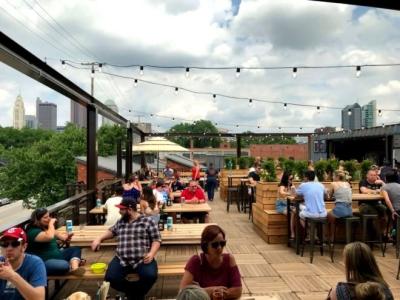 Rooftop patio with patrons seated at wooden picnic tables under string lights with skyline in background at BrewDog Franklinton