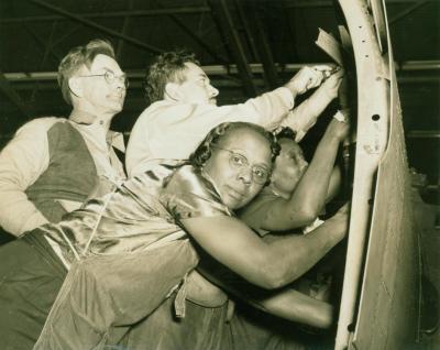 Two white men and two black women assemble an airplane part.