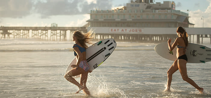 Two girls with surfboards run into the ocean by the Pier and Joe's Crab Shack.