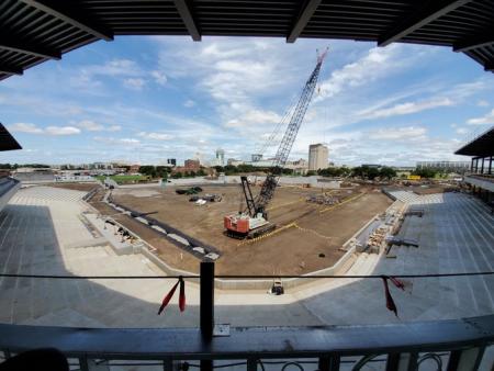 large crane overtop dirt field that will one day be baseball diamond, concrete structures under construction, blue sky overhead overlooking the Wichita skyline in background