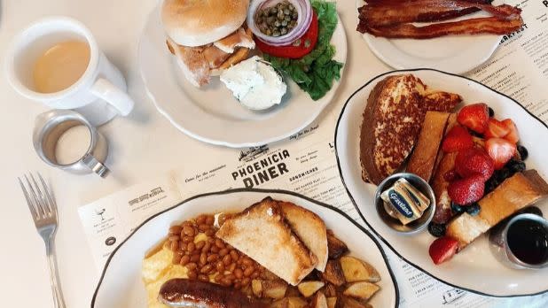 A photo of a spread of food from Phoenicia Diner: a burger, toast with baked beans and sausage, french toast with strawberries, bacon, and coffee