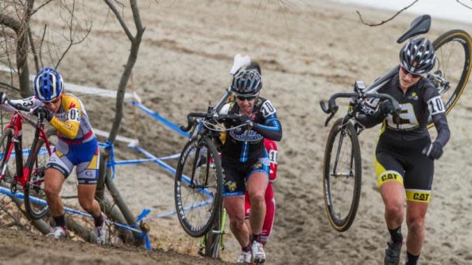 Three female bikers carrying their bikes up a muddy hill.
