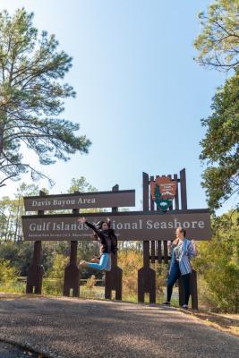 Two women posing outside in front of a sign surrounded by trees.