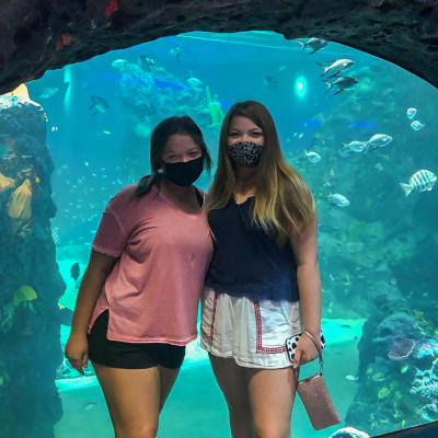 Two young ladies wearing masks and posing for a photo in front of an aquarium