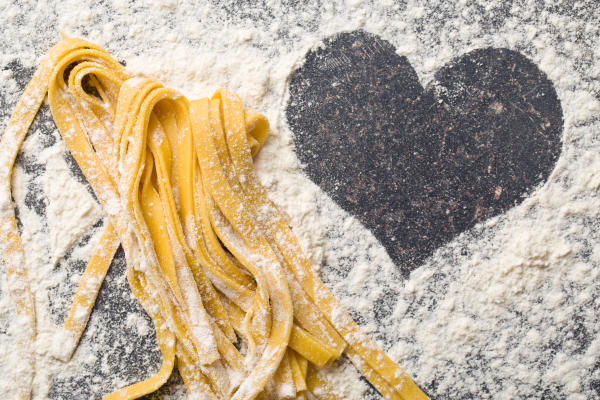 A pile of fresh-made pasta sits on a flour-dusted counter with a heart shaped clearing.