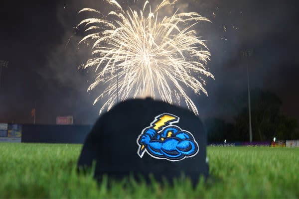 Baseball cap with Trenton Thunder Baseball team logo placed on grass of baseball field. Fireworks exploding in the distance.