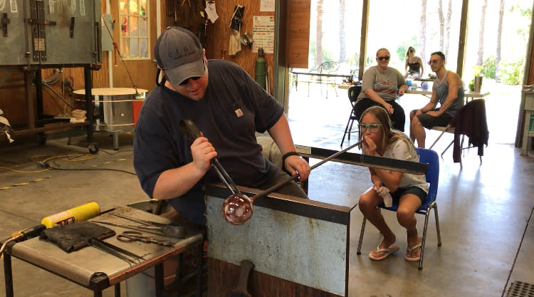 A young girl blows into a metal tube fitted with a glass ball at Orbix Hot Glass