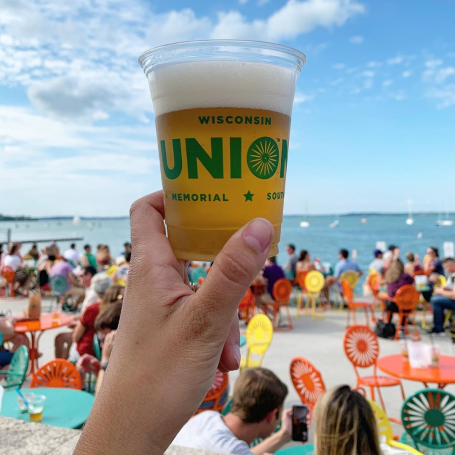 A person holds up a beer with the iconic Memorial Union Terrace chairs in the background