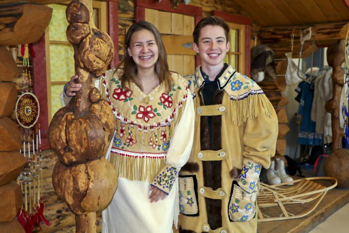 Alaska Native boy and girl dressed in traditional clothing standing next to a log cabin